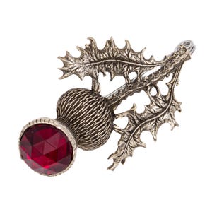 Puede incluir: Un broche de plata con forma de cardo con una gema roja en el centro. El cardo es un símbolo tradicional escocés.
