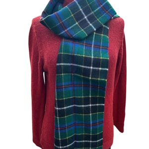 May include: A green, blue, and black plaid scarf with fringe is draped over a red sweater. The scarf features a classic tartan pattern with white and yellow accents. The scarf is made of a soft material and is suitable for cold weather.