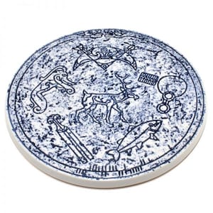 Può includere: Un sottobicchiere rotondo in ceramica bianca con un design blu di un cervo, un pesce e altri simboli. Il design ricorda l'arte nordica antica.