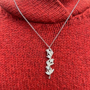 May include: A silver-colored necklace with a floral pendant. The pendant features a vertical arrangement of stylized flowers and leaves. The necklace is displayed against a red knitted sweater.