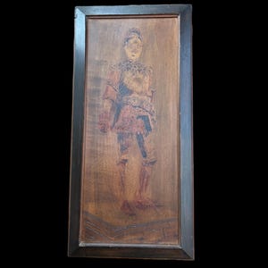 Op de afbeelding: Een ingelijst houtsnijwerk van een persoon in harnas. Het snijwerk is geschilderd in tinten bruin, rood en zwart. De lijst is van donkerbruin hout.