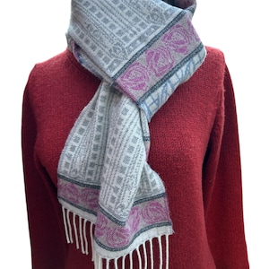 May include: A grey and white patterned scarf with pink accents and fringe. The scarf is wrapped around a red sweater.