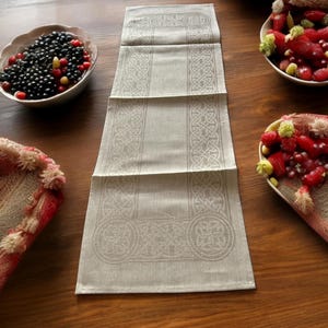 Puede incluir: Un camino de mesa de lino blanco con un diseño de nudo celta. El camino de mesa está sobre una mesa de madera con cuencos de fruta y bayas.