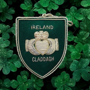 Könnte beinhalten: Ein grüner Filzschild mit goldener Stickerei, die das Claddagh-Symbol darstellt, ein Herz, das von zwei Händen gehalten wird, mit einer Krone oben. Der Schild trägt die in Gold gestickten Worte "IRELAND" und "CLADDAGH".