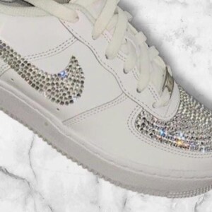 K&ouml;nnte beinhalten: Wei&szlig;e Leder-Sneaker mit niedrigem Schaft, einem mit Kristallen besetzten Nike-Swoosh und einer mit Kristallen besetzten Zehenkappe.