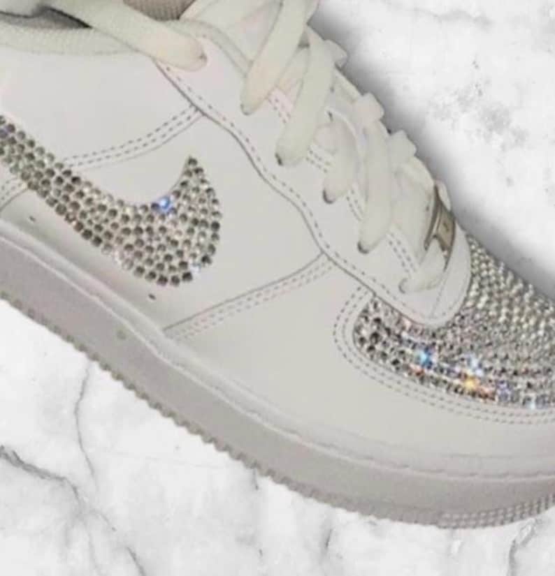 K&ouml;nnte beinhalten: Wei&szlig;e Leder-Sneaker mit niedrigem Schaft, einem mit Kristallen besetzten Nike-Swoosh und einer mit Kristallen besetzten Zehenkappe.