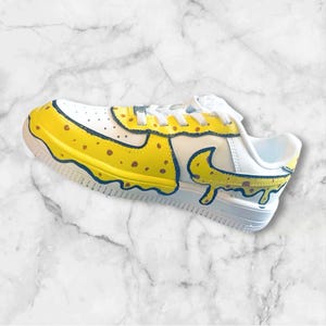 May include: White sneakers with a yellow and brown paint design. The paint is dripping down the sides of the shoe, creating a cartoon-like effect.