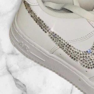 K&ouml;nnte beinhalten: Wei&szlig;e Lederschuhe mit einem mit Diamanten besetzten Nike-Swoosh.