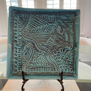 May include: A square, turquoise ceramic plate with a textured surface and dark brown etched designs. The plate is displayed on a decorative metal stand, resting on a vintage book. The plate's design includes geometric and organic shapes.