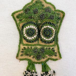 May include: A green and white beaded ornament with a star shape and fringe. The ornament has two circular designs with white and green beads.