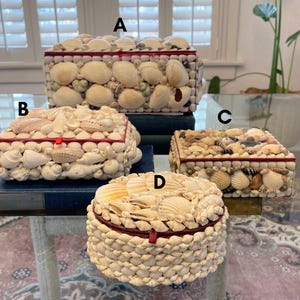 May include: Four decorative boxes made of seashells. The boxes are different sizes and shapes. The boxes are all white with red trim. The boxes are arranged on a blue surface.