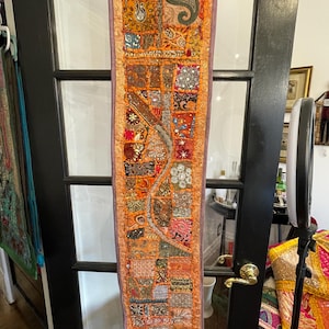 May include: A long, narrow tapestry with a patchwork design. The tapestry is made of various fabrics in shades of orange, brown, red, and purple. It is decorated with sequins, beads, and embroidery.
