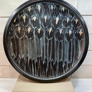 Peut inclure: Une assiette ronde en céramique avec un fond marron foncé et un bord noir. L'assiette présente un motif répété de fleurs stylisées noires en forme de tulipes avec des accents blancs et beiges. L'assiette est présentée sur un socle beige.