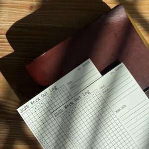 May include: Two 2026 Work Out Log sheets with a grid layout for tracking fitness goals. The sheets are white with black text and are partially covered by a dark brown leather notebook. The text "2026 Work Out Log" is visible.