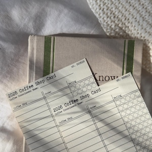 May include: Two "2026 Coffee Shop Card" sheets with lines for order and rating, and the text "Coffee Connoisseur Name" and "Coffee Shop". The cards are on top of a beige notebook with green stripes. The background is a white knitted blanket.