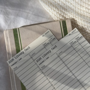 May include: Two "2026 Library Card" forms with lined spaces for author, title, and borrower's name, along with a book with green stripes. The forms have the number "000619" printed on them. The background includes a white knitted blanket.