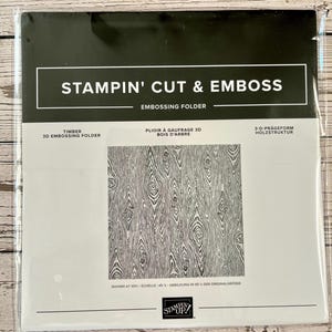 May include: A Stampin' Cut & Emboss 3D embossing folder in a clear plastic package. The packaging features the text Timber 3D Embossing Folder and displays a black and white wood grain pattern, with the Stampin' Up! logo at the bottom.