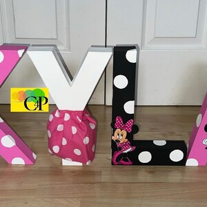 Op de afbeelding: Roze en zwarte letterdecoraties die de naam "SKYLAR" spellen. Elke letter is versierd met witte stippen en een Minnie Mouse-personage.
