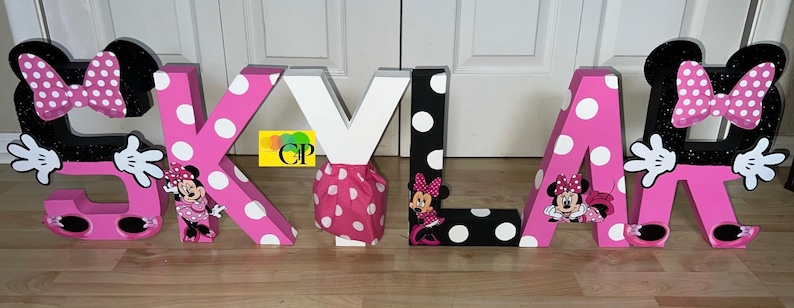 Op de afbeelding: Roze en zwarte letterdecoraties die de naam "SKYLAR" spellen. Elke letter is versierd met witte stippen en een Minnie Mouse-personage.