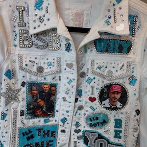 Op de afbeelding: Een wit spijkerjack dat rijkelijk is versierd met strass-steentjes, blauwe glitteraccenten en patches. Het bevat de teksten "I BSB", "WHY", "I'LL BE THE ONE", "IT'S GOTTA BE YOU" en "AJ", samen met een foto van een boyband en een portret van een man.