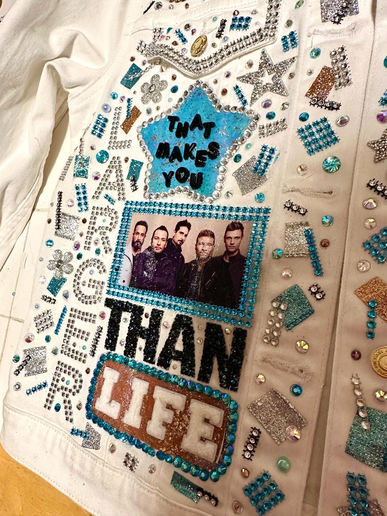 Close-up of custom jacket with rhinestones, teal star patch, band photo, and text reading That Makes You Larger Than Life.