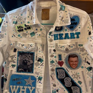 Op de afbeelding: Een wit spijkerjack versierd met blauwe en zilveren strass-steentjes, pailletten en patches. Het jack bevat een foto van een boyband, de teksten "I'll be the one", "My heart", "Tell me why" en "BSB", en accenten in de vorm van sterren en bloemen.