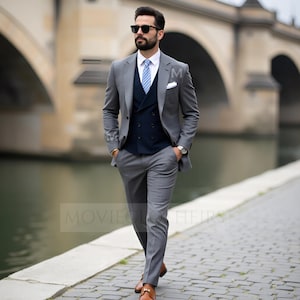May include: A three-piece gray suit with a navy blue vest, white shirt, and blue striped tie. The suit is worn with brown leather shoes and sunglasses. The person is walking on a cobblestone path near a body of water.