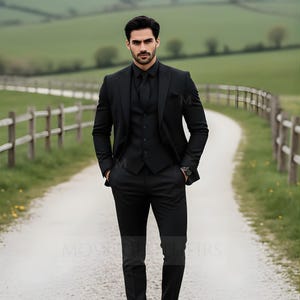 May include: A black three-piece suit, including a jacket, vest, and trousers, is worn by a person standing on a gravel path. The suit is paired with a black dress shirt and tie. A watch is visible on the person's wrist. The background features a green field and a wooden fence.