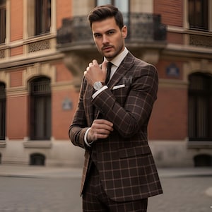 May include: A brown plaid suit with a white dress shirt and a brown tie. The suit has a classic cut and is worn with a wristwatch. The background features a brick building.