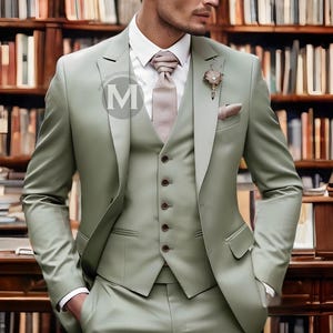 Puede incluir: Un traje verde salvia de tres piezas con una corbata de estampado rosa claro y un pequeño alfiler de flor. El traje lo lleva una persona de pie frente a una estantería.