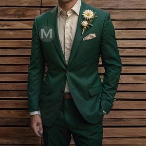 May include: A dark green suit jacket with a white button-down shirt underneath. The jacket has a single button closure and a pocket square in the left breast pocket. A small white flower boutonniere is pinned to the lapel.
