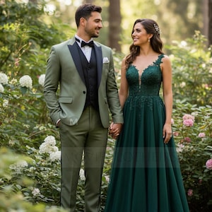 Peut inclure: Un couple en tenue de cérémonie, posant à l'extérieur. L'homme porte un costume vert sauge avec un gilet noir et un nœud papillon. La femme porte une robe vert foncé avec des détails en dentelle. Ils se tiennent la main, entourés de verdure.
