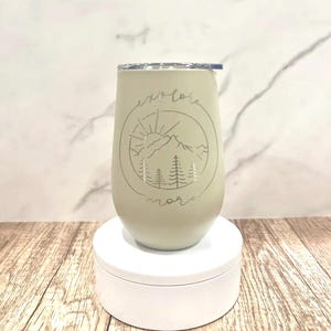 May include: A light green stainless steel tumbler with a white lid. The tumbler has a graphic of a mountain range with a sun and trees. The text "explore more" is written around the graphic.