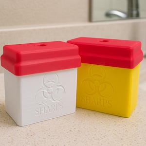 May include: Two rectangular sharps containers, one white and one yellow, each with a red lid. The containers have a biohazard symbol and the word "SHARPS" printed on them. They are sitting on a speckled countertop.