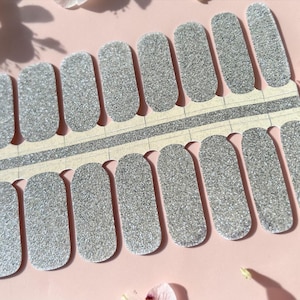 May include: Silver glitter nail polish strips arranged on a white backing. The nail strips are in various sizes and shapes, with a sparkly, textured appearance. The background is a soft pink with white flowers.