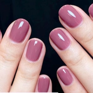 May include: Close-up of hands with polished nails. The nails are painted a solid mauve color with a glossy finish. The oval-shaped nails have a reflective sheen, and the skin tone is fair. The background is a dark, solid color.