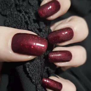 May include: A hand with polished nails in a close-up shot. The nails are painted a deep burgundy with a glittery finish. The hand is positioned near a black, textured fabric, creating a contrast between the smooth nails and the rough fabric.