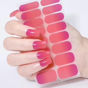 May include: A hand holding a sheet of pink and red ombre nail wraps. The nails on the hand are painted with a matching pink ombre design. The nail wraps are in an oval shape.