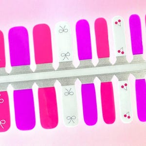 May include: Nail art strips featuring a variety of designs. The strips include solid magenta and pink colors, white strips with small bows, and white strips with red cherries. The strips are on a silver backing.