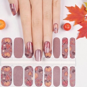 May include: Nail art strips with autumn-themed designs. The strips feature pumpkins, leaves, and acorns in shades of orange, red, and brown on a mauve background. The image also includes a hand with matching nail art and autumn foliage.