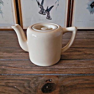 Peut inclure: Une petite théière en céramique de couleur crème avec un couvercle. La théière a un corps arrondi et un bec verseur sur le côté. La théière est posée sur une surface en bois.