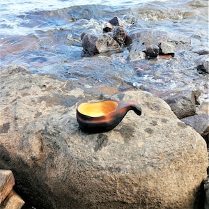 May include: A dark brown wooden bowl with a yellow interior, sitting on a gray rock near a body of water.