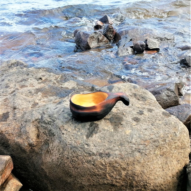 May include: A dark brown wooden bowl with a yellow interior, sitting on a gray rock near a body of water.