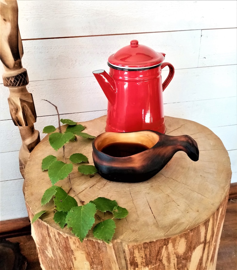 May include: A red enamel teapot and a wooden cup with a dark brown liquid inside, sitting on a wooden stump with green leaves.