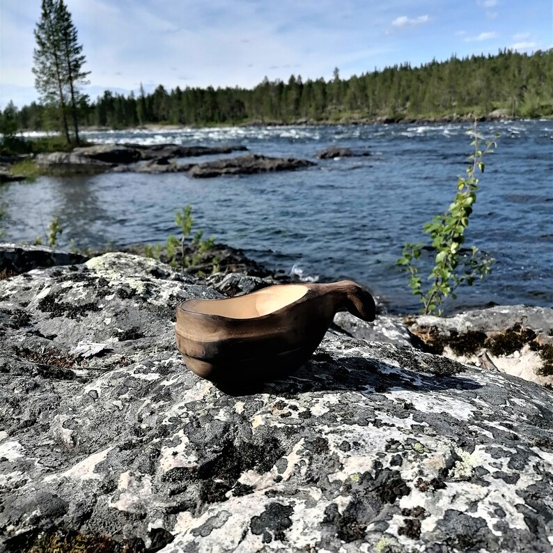 May include: A dark brown wooden bowl with a handle, sitting on a gray and white rocky surface near a body of water. The bowl is made from a single piece of wood and has a natural, rustic look.