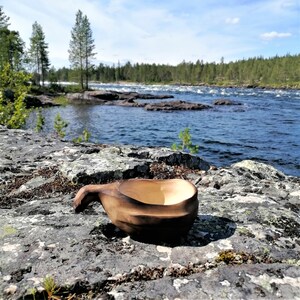 May include: A wooden cup with a handle sits on a rocky surface near a river. The cup is dark brown and has a natural wood grain. The river is blue and there are trees in the background.
