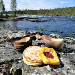 May include: Two wooden bowls and two pastries on a rocky surface by a river. The pastries are a round, flat bread and a pastry with a red filling. The river is in the background and there are trees on the far side.