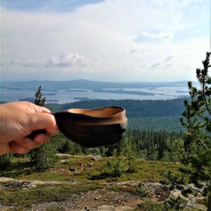 May include: A wooden bowl with a dark brown finish is held in a hand against a backdrop of a scenic landscape. The landscape features a vast body of water with numerous islands and a forested hillside.
