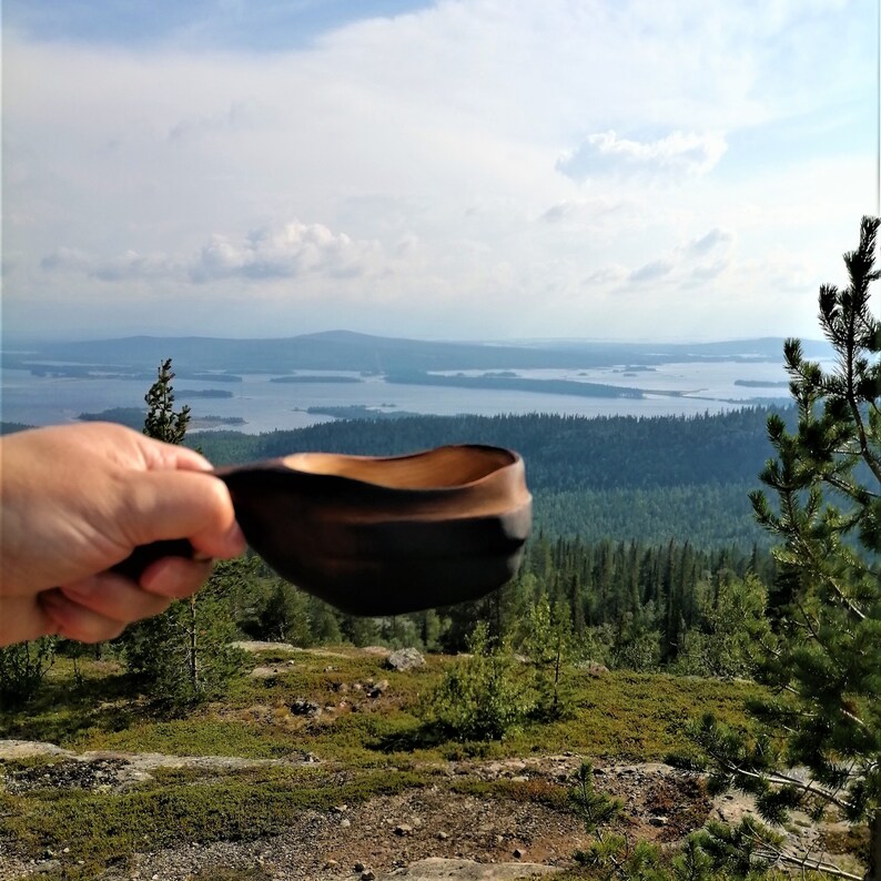 May include: A wooden bowl with a dark brown finish is held in a hand against a backdrop of a scenic landscape. The landscape features a vast body of water with numerous islands and a forested hillside.