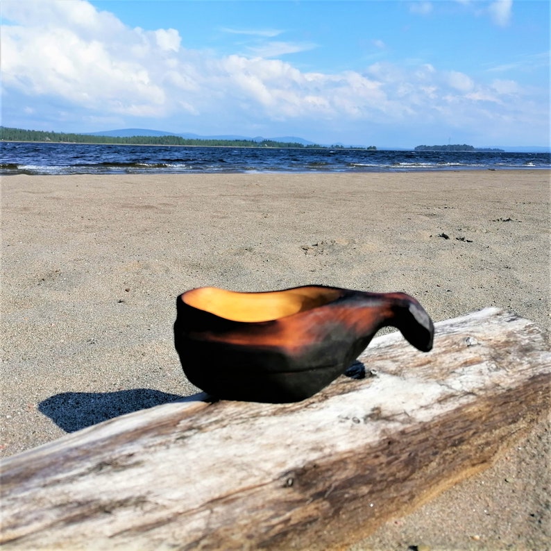 May include: A wooden bowl with a dark brown exterior and a light brown interior sits on a weathered log. The bowl has a handle and is resting on a sandy beach with a lake in the background.